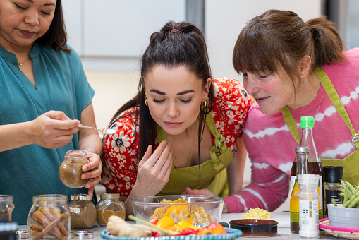 Group of mixed ethnicity and age people participating in a Thai cooking class together. They are standing in a kitchen wearing aprons, learning a new skill in the North East of England. The women are bending over to smell the ingredients.