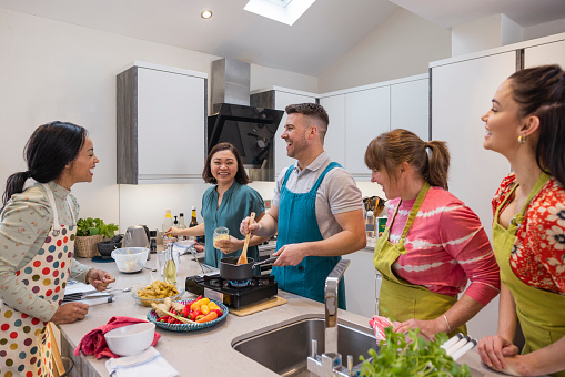 Group of mixed ethnicity and age people participating in a Thai cooking class together. They are standing in a kitchen wearing aprons, learning a new skill in the North East of England. They are making a massaman beef curry, adding coconut milk to a pan.