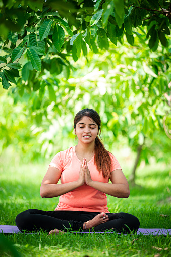 Outdoor image of Young Asian/ Indian woman sitting in Namaste or prayer yoga position and relaxing at park. Concept of Yoga and healthy lifestyle. Concept of Healthy lifestyle and yoga at park.