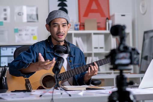 Young Asian male graphic designer blogger influencer playing guitar talking looking at camera shooting education tutorial vlog training filming video course for social media at studio.