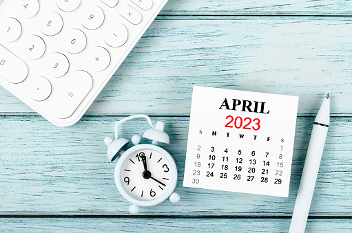 April 2023 Monthly calendar year and alarm clock with calculator on blue wooden background.
