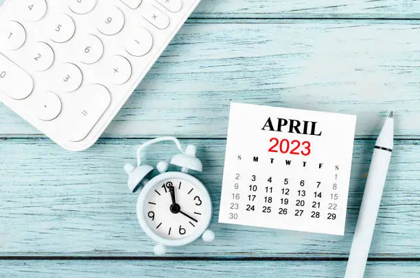April 2023 Monthly calendar year and alarm clock with calculator on blue wooden background.