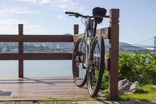 Light green bicycle resting on a brown wooden deck overlooking the sea. There is green grass on the side and a distant bridge on the background.