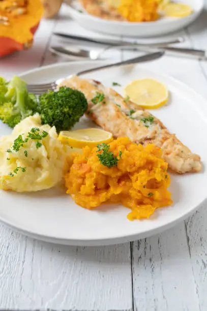 Pan fried natural white fish fillet with broccoli, mashed potatoes and red kuri squash puree. Served on a plate on bright and white wooden background with space for text