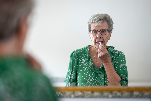 A senior woman getting ready for a day out, she is putting lip balm on her lips while looking in a mirror at home in Newcastle Upon Tyne, England.