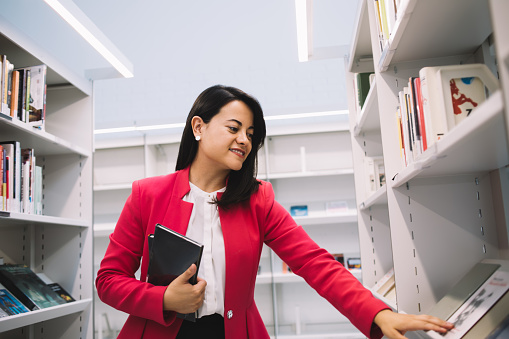 Black haired ethnic lady in formal clothes standing between bookshelves in bright library and looking at book while picking one