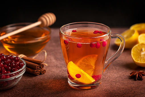 Herbal natural vitamin hot tea made with sour sliced lemon citrus, cranberries and sweet honey served in transparent glass cup on dark brown table with ingredients, cinnamon and anise as healthy drink