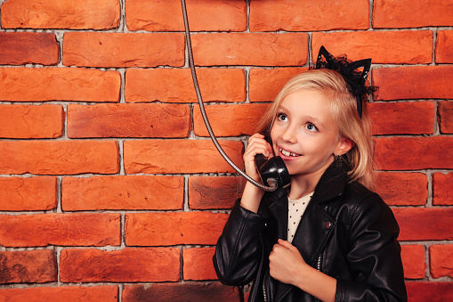 Portrait little girl model in leather jacket calling old payphone at brick wall background, smiling. Child emotional lady talking telephone outdoor. Communication lifestyle concept. Copy text space