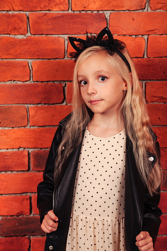 Portrait fashionable blonde girl in leather jacket standing at brick wall background, looking at camera. Little model with positive emotion, smiling. Fashion style concept. Copy text space
