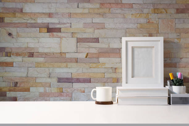 istock Stylish workplace blank picture frame coffee cup, stationery and books on white table against brick wall. 1454408713