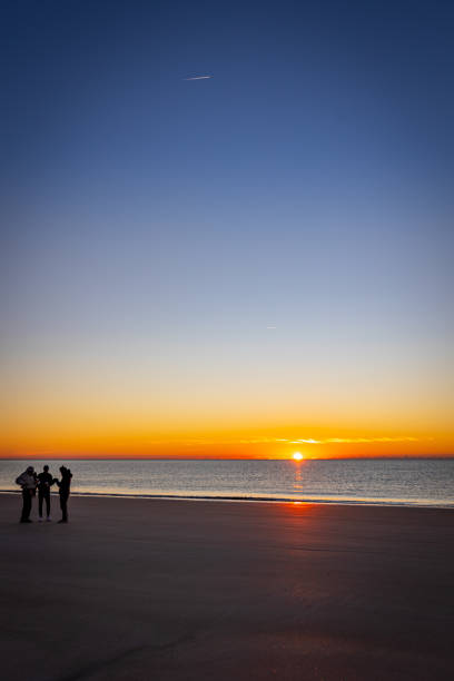 pessoas na praia durante o nascer do sol de cor dourada - silhouette three people beach horizon - fotografias e filmes do acervo