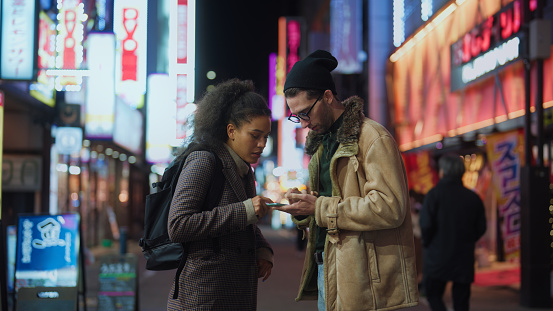 Two tourists are using a smart phone to explore the town in Tokyo at night.