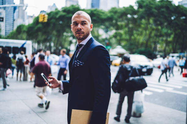 Side view of reserved businessman in navy blue suit standing with folder and smartphone while looking away on blurred background with people walking on sidewalk