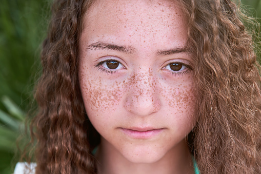Close-up portrait of a teenage girl with freckles on her face and curly long hair. Looking into the camera. Close up of face. Natural beauty, skin care, cosmetology concept