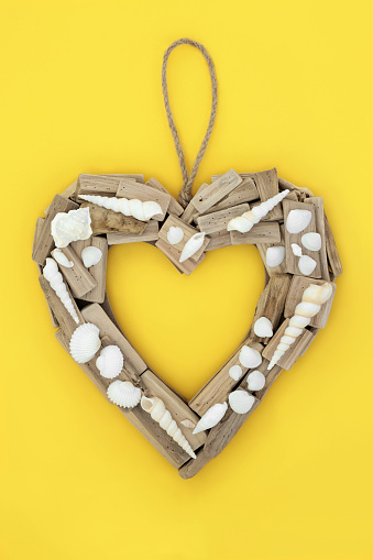 Driftwood heart shaped wreath with seashells. Natural rustic beautiful holiday and travel destinations object. Symbol for Mothers Day, anniversary, birthday, Valentines Day. On yellow background.