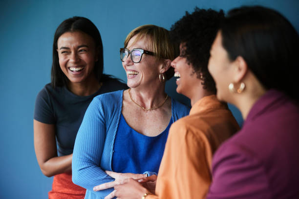 Diverse group of businesswomen laughing against a blue background Group of diverse businesswomen laughing while standing together in front of a blue background mixed age range stock pictures, royalty-free photos & images