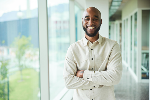 Portrait of a businessman smiling while standing with his arms crossed by windows in the corridor of a modern office