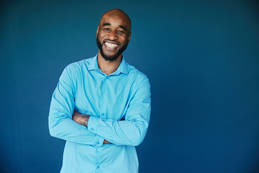 Portrait of a laughing businessman with a shaved head and beard standing with his arms crossed in front of a blue background