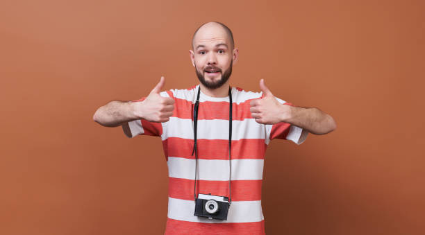 Charming young guy with a camera shows a like sign with his hands. stock photo