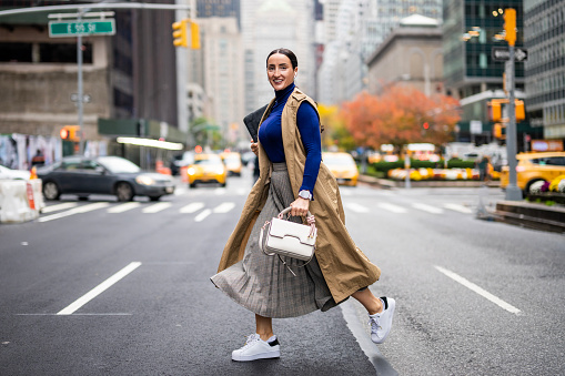 Beautiful woman in a beige sleeveless coat, skirt, turtleneck sweater, white sneakers and with a handbag seen crossing a street in Manhattan, New York, while using her wireless headphones and talking on the phone during a break at work.