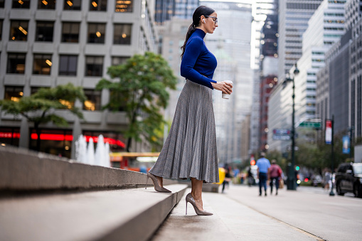Businesswoman with eyeglasses and wireless headphones wearing a turtleneck sweater, pleated skirt and high heels seen holding a cup while walking down the stairs in front of the building in Manhattan during a break at work.