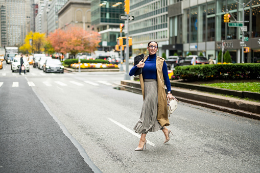 Fashionable businesswoman in high heels, beige sleeveless coat, skirt and turtleneck sweater with a handbag and a laptop seen crossing a street in Manhattan, New York, after finishing work.