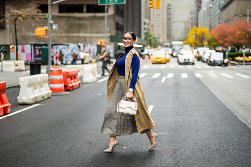 Fashionable businesswoman in high heels, beige sleeveless coat, skirt and turtleneck sweater with a handbag and a laptop seen wearing eyeglasses and crossing a street in Manhattan, New York, after finishing work.