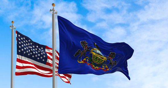 Pennsylvania state flag waving alongside the national flag of the United States on a sunny day. The flag of Pennsylvania features a coat of arms on a blue field. Realistic 3d illustration
