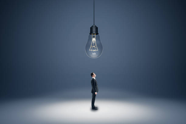 istock Innovation, solution and creativity concept with businessman in black suit looking up at the ceiling with a big hanging light bulb on abstract empty grey area background 1454440140