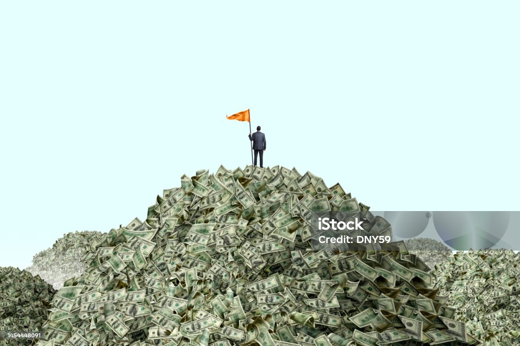 Man Planting Flag On Piles Of Cash A businessman stands at the top of a mountain of money as he holds a large orange flag attached to a pole. He stands with his back to the camera as he looks out into the distance towards other mountains of money that he has yet to conquer. Currency Stock Photo
