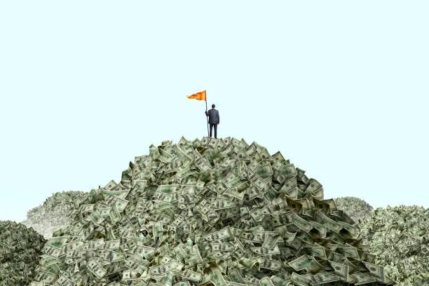 A businessman stands at the top of a mountain of money as he holds a large orange flag attached to a pole. He stands with his back to the camera as he looks out into the distance towards other mountains of money that he has yet to conquer.