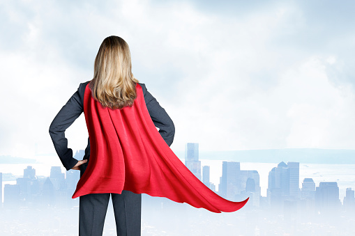 A rear view of a businesswoman wearing a red cape as she looks out toward the skyline of a large city.