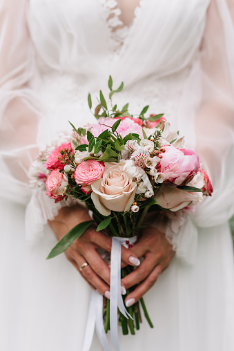 wedding bouquet of white and pink roses and peonies in the hands of the bride on the background of a white dress