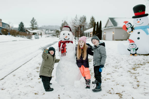 Elementary age twin brother and sister pose with their younger cousin beside a snowman they built together in the front yard of their home while on Christmas vacation.