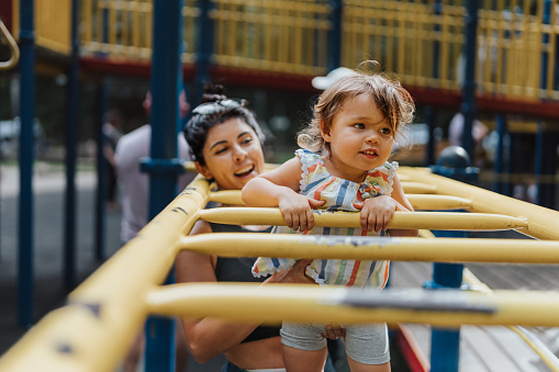 A cute multiracial toddler girl has fun using the monkey bars at the playground with her Latin American nanny's help.
