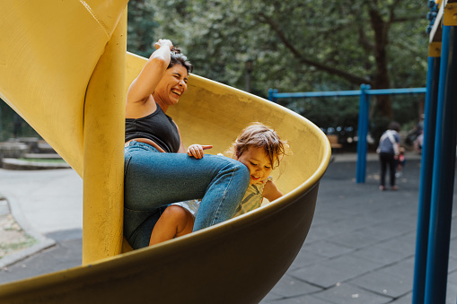 A beautiful Latin American woman laughs while riding down a spiral yellow slide with a cute two year old toddler girl.