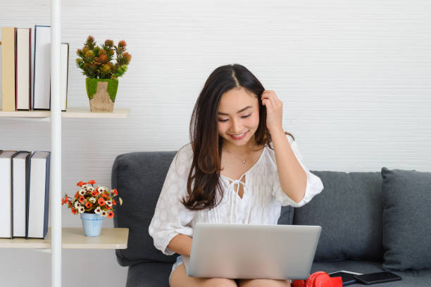 istock Beautiful young Asian woman using laptop doing work online study while sitting on couch at home. shopping or chatting online in social network. 1454491807