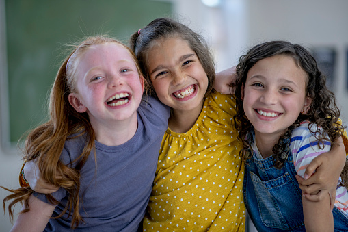 A small group of three elementary school girls huddle in closely together with their arms around one another as they pose for a portrait.  They are each dressed casually and have smiles on their faces.