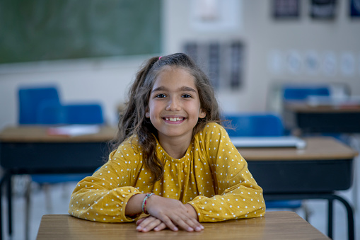 A young female Elementary student sits at her desk as she poses for a portrait.  She  is dressed casually and has a smile on her face.