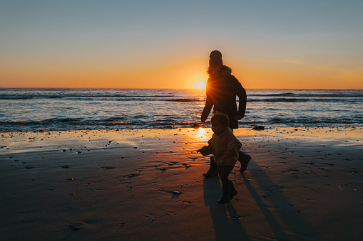 Silhouette of a multiracial woman playfully chasing her two year old daughter along a sandy beach in the Pacific Northwest region of the United States as the sun sets over the ocean on a cool winter evening.