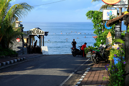 View of the sea with many surfers in the waves at Batu Bolong Beach in Canggu, Bali, Indonesia. The road ends at the beach and several cafes and restaurants line the road including the Sandbar and others.