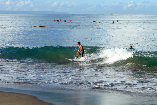 A surfer riding a beach break wave at Batu Bolong Beach in Canggu, Bali, Indonesia with other surfers waiting for waves in the background. Batu Bolong is famous for riding long boards in Bali.