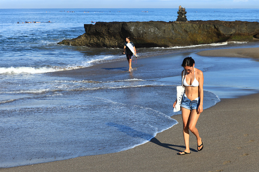 An Asian woman walking along the beach at Batu Bolong Beach in Canggu, Bali, Indonesia, wearing sunglasses while behind her a surfer is walking with is surfboard.