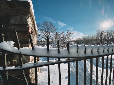 Iron fence with snow stuck on top and a beautiful Michigan landscape view peeking out from over the fence, taken from the road and drive leading into a Cemetery in Faithorn Township in The Upper Peninsula during a Polar Vortex winter storm. Sun and beautiful daytime skies above.