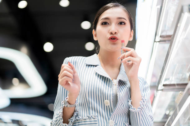 femme asiatique shopping dans le magasin cosmétiques, bonheur souriant femme asiatique faire drôle face à la caméra tout en utilisant à la main goûteur lipstique au magasin de cosmétiques dans le centre commercial department mall - asian ethnicity women shopping mall perfume photos et images de collection