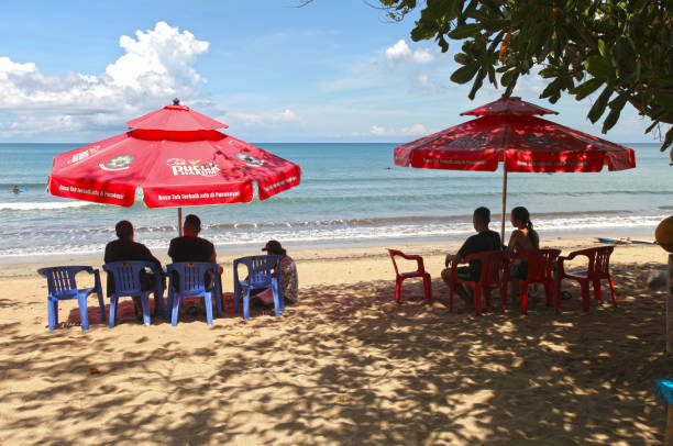 kuta beach à bali, indonésie avec un beau ciel bleu par une chaude journée ensoleillée et des parasols ou des parasols. des gens assis sur des chaises en plastique dans l’ombre sous des parasols. - parasol umbrella asian ethnicity asian culture photos et images de collection