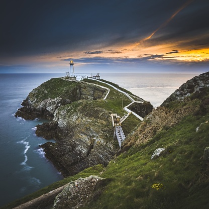 An aerial shot of South Stack Lighthouse on a cliff under the dramatic clouds and golden sunset