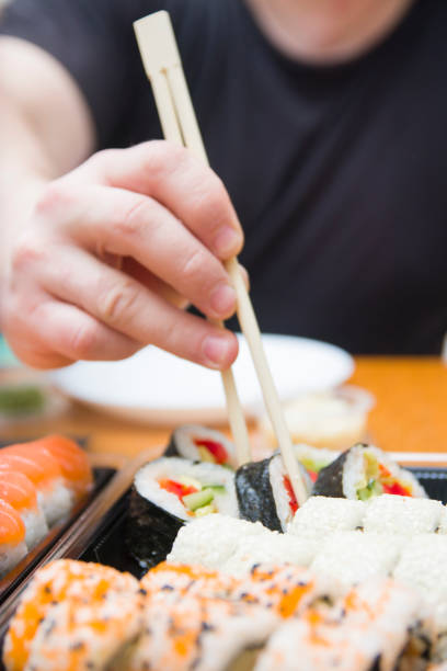 istock Close-up high-angle view hands of unrecognizable young man holding sushi with chopsticks sitting at desk. Closeup of male enjoying sushi. 1454520710