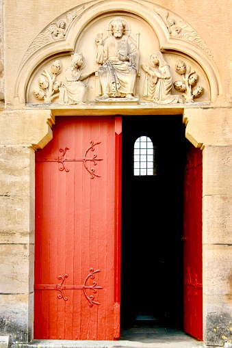 Vertical closeup photo of a vibrant red door, half open, showing a small arched window inside the black interior of an ancient stone church with carved sculptures above the doorway. Burgundy, France.
