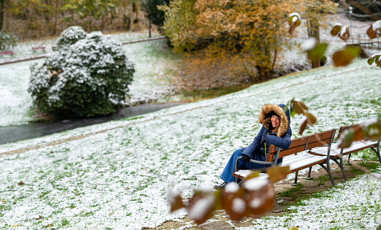 Lady looks back to the camera posing while she is in a park outside sitting on a bench during a snowy day. She looks happy and comfortable
